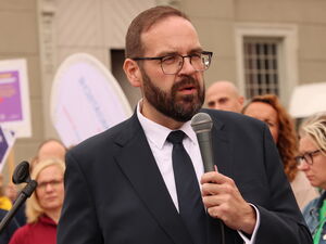 Mann in Anzug und Krawatte spricht in ein Mikrofon. Dahinter Menschen mit Plakaten und Fahnen.