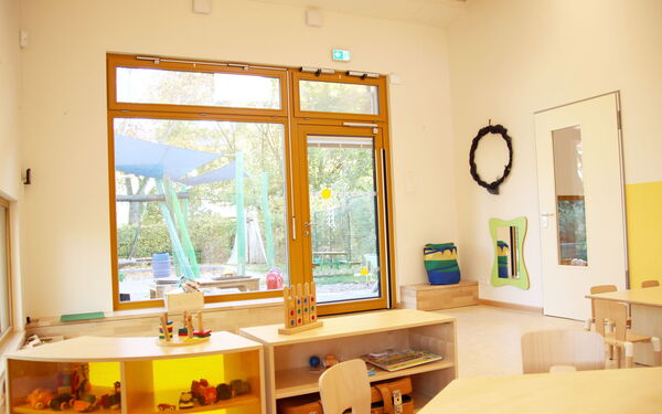 Spielraum. Großes Fenster, Terrassentür, die zu einem kleinen Spielplatz führt. Im Raum, zwei kleine Regale mit Spielzeug, zwei kleine Tische mit Stühlen.