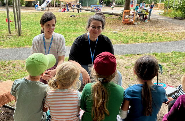 4 Kinder sitzen auf einer Bank von hinten fotografiert. Vor ihnen sitzen zwei Frauen, Im Hintergrund ein Spielplatz.