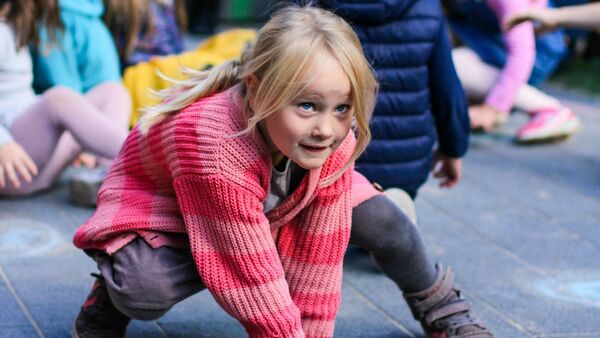 Ein Mädchen tanzt auf dem Boden. Mädchen hat lange blonde Haare, eine pinke Strickjacke und eine graue Hose an. Hinter ihr sitze Kinder auf dem Boden.