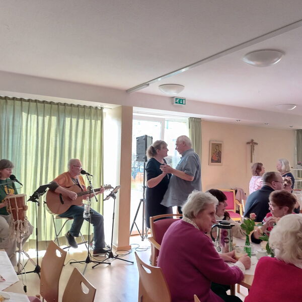 Heller Raum mit großen Fenstern. Kaffeetafeln und zwei Menschen die am Fenster sitzen und Musik machen. Rechts von dem Musikern ein tanzendes Paar.