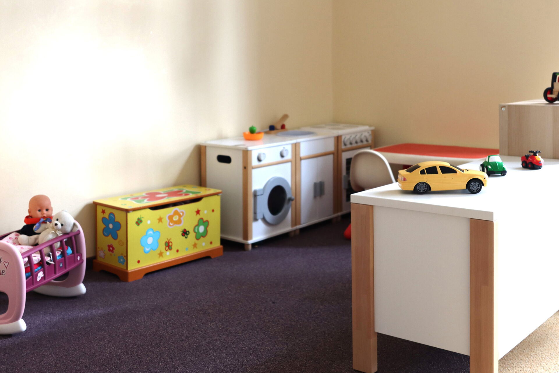 In der Zimmerecke ein Kinderherd, Spüle und Waschmaschine. Daneben eine gelbe Truhe mit bunten Blumen drauf.