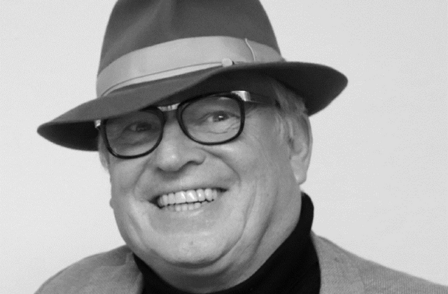 Schwarz-Weiß Portrait: Mann mit Brille und Hut lächelt breit.  Mann ist alt, graue Haare. Volles Gesicht.