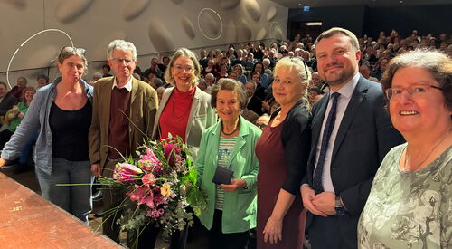 Preisverleihung: Gruppenbild vor der Bühne stehen 7 Menschen. Das Publikum hinter ihnen. Frau in der Mitte hält großen Blumenstrauß.