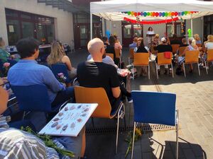 Ein weißer Pavillon, bunt geschmückt mit Luftballons und Luftschlagen. Darunter Menschen die auf Stühlen sitzen. Vorn steht eine Rednerin an einem Pult.