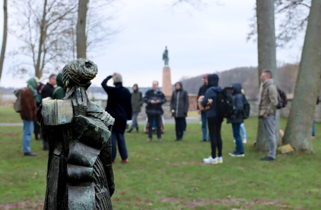 Im Vordergrund ein Denkmal. Dahinter draußen auf einer Wiese zwischen Bäumen stehen einige Menschen in einem Kreis.