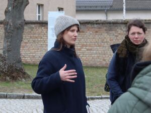 Draußen. Zwei Frauen in Winterkleidung. Frau mit grauer Mütze und dunkelblauer Jacke erklärt etwas. Sie hält die Hand auf Brusthöhe.