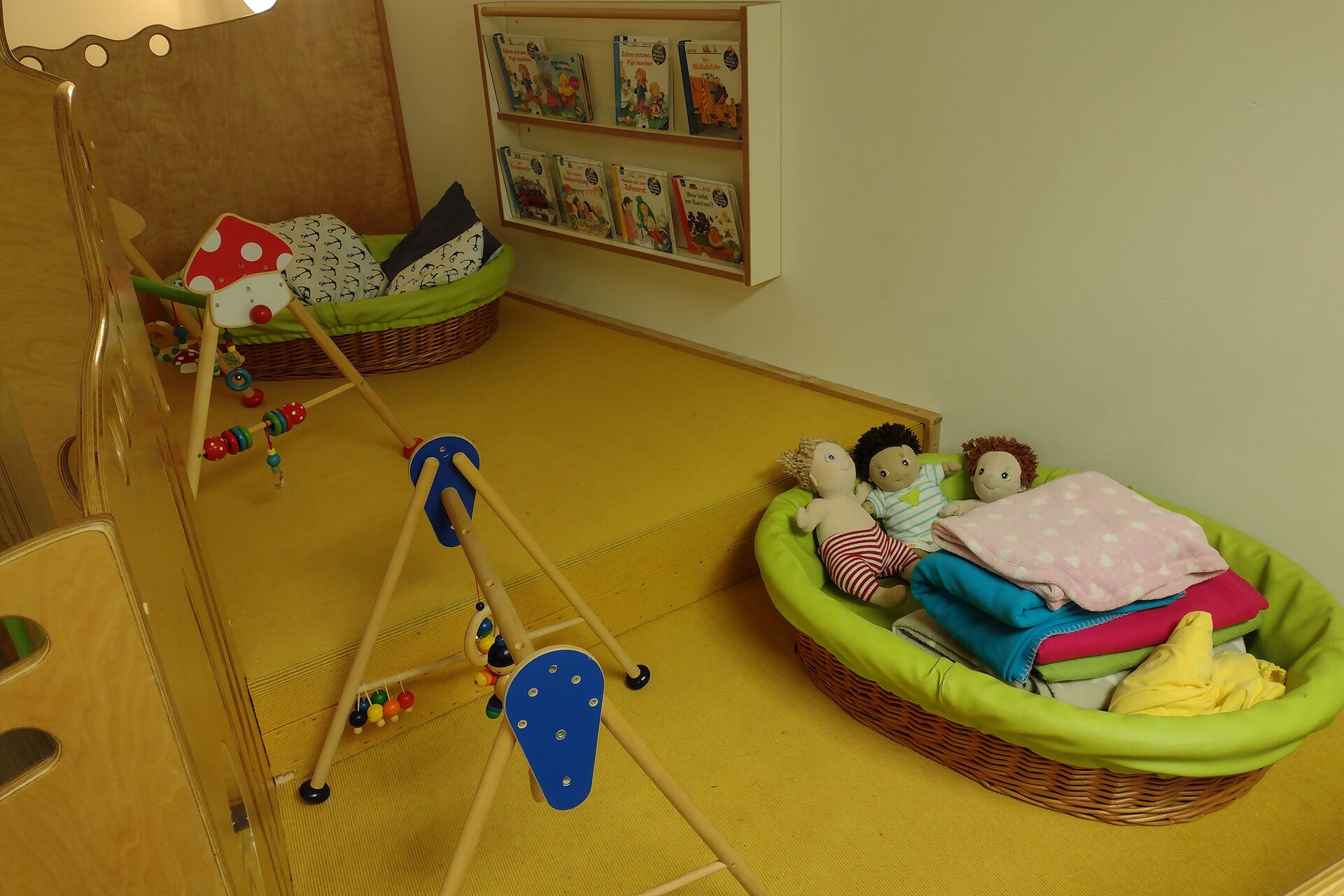 Zwei Kleinkindkuschelnester mit Decken und Puppen als Inhalt und zwei Spielbögen auf gelbem Teppich