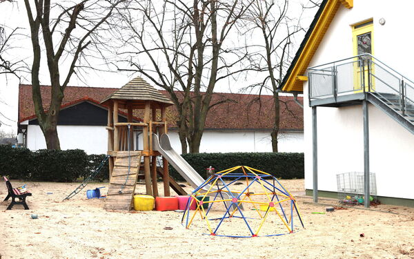Spielplatz mit Krettergerüst mit Rutsche und einer Bank. Umgeben von einer Hecke. Daneben weißes Haus mit einer Außentreppe die zu einer gelben Tür im Obergeschoss des Hauses führt.