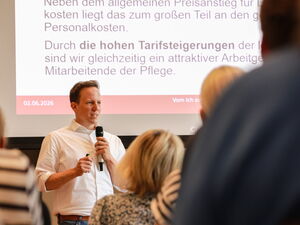 Teilnehmende von hinten. Vorne ein Mann im weißen Hemd mit Mikrofon. Dahinter läuft eine Präsentation auf großer Leinwand über Tariflohn.
