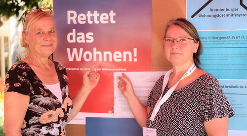 Zwei Frauen stehen vor einer Info-Tafel. An der Tafel sind Informationen zum Thema wohnen vom Brandenburger Wohnungslosenhilfesystem. Beide zeigen auf ein Plakat Text: Rettet das Wohnen!