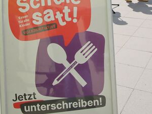 Draußen, Sonne scheint. Aufsteller Schild Text: Schule satt! Essen für alle Kinder. Jetzt unterschreiben.       Dahinter drei Personen. 2 Frauen, ein Mann in der Mitte am Mikro.