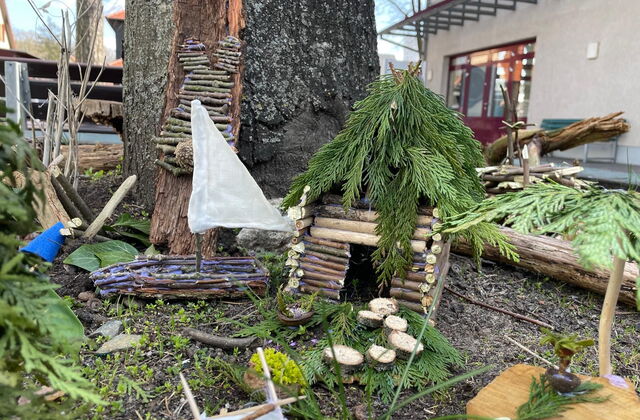 An einem Baum, liegen gebastelte Gegenstände aus Holz und anderen Naturmaterialien. Ein kleines Segelboot, eine Art Hütte mit Tannenzweigen-Dach.