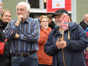Mehrere Menschen stehen neben- oder voreinander. Sie haben Babyrasseln oder Protestschilder in der Hand oder Trillerpfeifen im Mund.