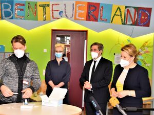 Ein farbenfroher Raum, grüne Wand. Vier Personen mit FFP 2 und OP Masken stehen um einen runden Stehtisch. Darauf eine Packung mit Corona Tests. Darüber bunte Buchstaben auf farbigen Papier: ABENTEUERLAND