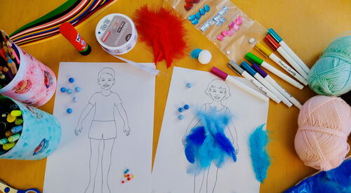 Tisch von oben mit Mal- und Bastelutensilien. Zwei Bilder nebeneinander, Junge und Mädchen. Auf dem Bild des Mädchens sind blaue Federn geklebt. Stifte, Schere, Wollknäuel, blaue Perlen, Prittstift