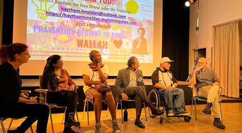 6 Menschen sitzen nebeneinander im Podiumsgespräch. Zwei Frauen, ein schwarzer Mann, ein ältere Mann im grauen Anzug, ein Rollstuhlfahrer im Aktivrollstuhl mit Basecap, Hoody und Jeans und ein Mann mit Glatze, Brille der in ein Mikro spricht.