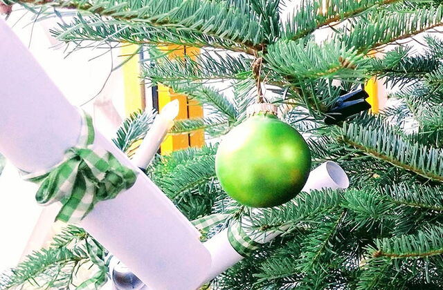 Tannenbaum mit grünen, roten Kugeln und Lichterkette geschmückt. An den zweigen hängen auch gerollte Zettel mit Schleifen drum.
