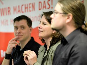 3 Personen sitzen nebeneinander. Von nahen und von der Seite fotografiert. Junger Mann, mit kurzen schwarzen Haaren, in der Mitte Frau mit dunkelbraunem schulterlangem Haar und junger Mann mit zurückgekämmten Haaren zu einem Zopf zusammengebunden mit Bril
