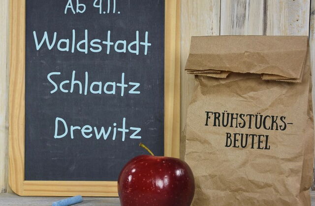 Eine Tafel, an der mit blauer Kreide geschrieben steht: Ab 9.11. Waldstadt Schlaatz Drewitz - Das Kreidestück liegt vor der Tafel sowie ein roter Apfel. Daneben ist eine gefüllte Papiertüte darauf steht: Frühstücksbeutel