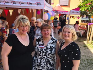 Gruppenfoto: Draußen, 3 Frauen in Sommerkleidern stehen vor bunt geschmückten Pavillons.