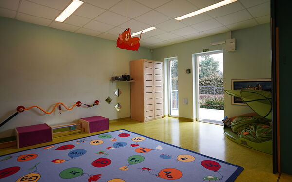 Raum mit Terrassantür. Im Raum liegt ein großer Kinderteppich mit bunten Luftballons auf denen Buchstaben stehen.