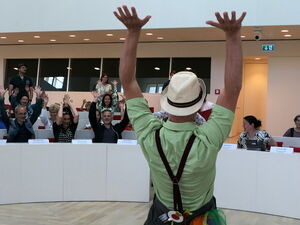Klinikclowns sorgen für gute Laune im weißen Plenarsaal. 2 Clowns strecken die Arme nach Oben, Kinder und Erwachsene auf den Sitzplätzen machen mit.