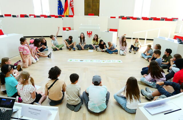 Kinder sitzen in einem großen Kreis auf dem Fußboden des Plenarsaal. In der Mitte liegen Bilder.