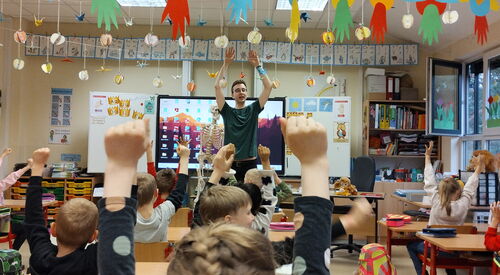 In einem Klassenraum. Ein Erwachsener steht vorne vor den Kindern an ihren Schulbänken. Alle strecken die Arme nach oben.