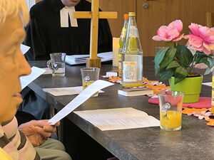 Ein Tisch. Am Kopfende sitzt der Pfarrer, ein Holzkreuz steht vor ihm. Am Tisch sitzen Senioren.