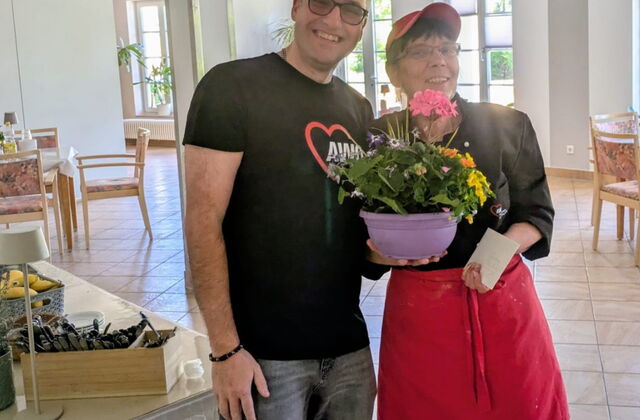 Cafeteria: Mann und Frau stehen im Zentrum und lächeln in die Kamera. Die Frau hält einee lila Blumenschale in der Hand.