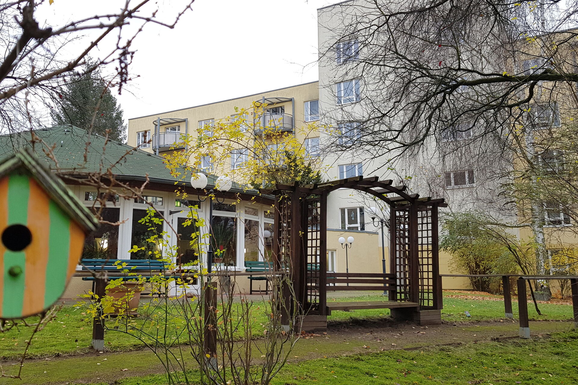 Parkanlage mit kleinem Häuschen mit grünem Dach. Vorne links ist ein kleines Vogelhäuschen. Im Hintergrund ein großes Wohnhaus.