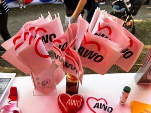 Ein Tisch. Auf dem Tisch: ein AWO-Schlüsselband, Eine Hand und ein Herz aus Ton in Rot mit AWO-Logo, ganz viele weiße AWO-Fähnchen stecken in einem Glas.