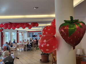 Großer Raum mit roten Luftballons mit weißen Punkten geschmückt. Senioren sitzen an Tischen beim Essen.