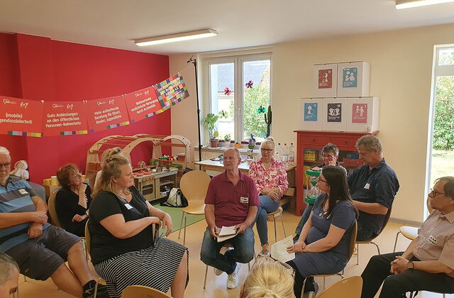 Raum mit großen Fenstern, 11 Menschen sitzen auf Stühlen, die in Kreisen angeordnet sind. In der Mitte sitzen sich zwei Frauen gegenüber ein Mann dazwischen.