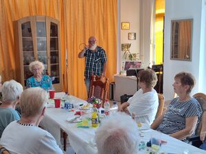 5 ältere Frauen an einer langen Tafel. Getränke stehen auf dem Tisch. Nähe des Kopfendes steht ein alte Mann mit weißem Bart, Brille und einem Mikrofon in der Hand.