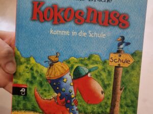 Eine Hand hält ein Buch in die Kamera. Buch: Der kleine Drache Kokosnuss kommt in die Schule       Ein kleiner Drache mit Schultüte