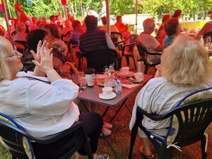 Menschen sitzen an kleinen Gartentischen bei Kaffee und Kuchen und klatschen.