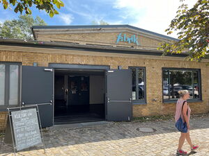 Eingang fabrik Potsdam. Beide Türen sind weit geöffnet. Der Schriftzug "fabrik" ist hellblau und miitig oben am Gebäude angebracht. Das Gebäude hat eine noch heute ansprechende und zeitgemäß wirkende fein gegliederte neoklassizistische Fassade.