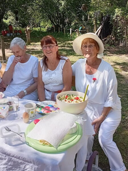 Drei Frauen in weißer Kleidung, die Frau ganz rechts mit Sonnenhut. Vor ihnen ein Picknicktisch mit einer großen Salat Schüssel.