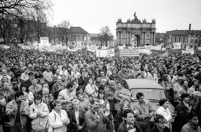Schwarz-Weiß Foto: Vor dem Brandenburger Tor in Potsdam stehen dicht gedrängt ganz viele Menschen. Einige von ihnen halten Plakate hoch, auf denen sie Freiheit oder das Absetzen der Regierung fordern.