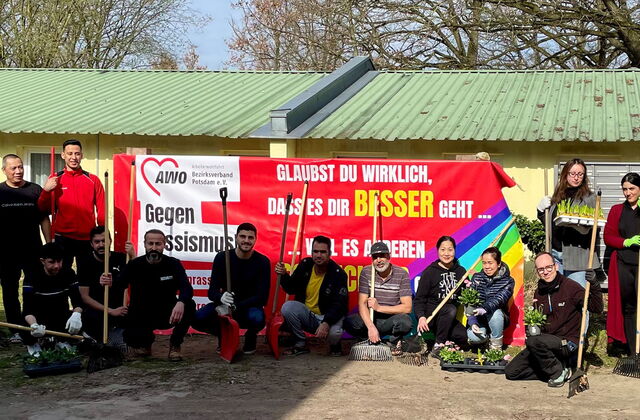 15 Menschen unterschiedler Nationen mit Pflanzen und Gartengeräten, Besen, Harke und Schaufeln. Die Menschen stehen und hocken vor dem AWO Banner