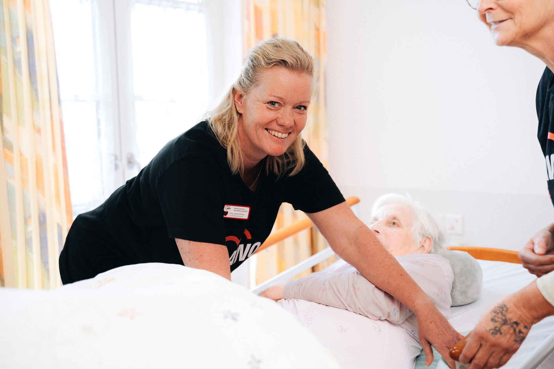 Eine Frau mit einem schwarzen AWO T-Shirt kümmert sich um eine alte Frau in einem Pflegebett und lächelt in die Kamera.