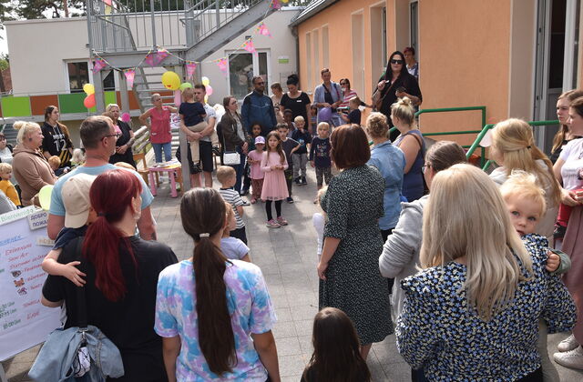 Viele Menschen / Kinder stehen neben einem Gebäude. Sie stehen in einem großen Kreis. Mütter mit kleinen Kindern auf dem Arm. Eine Treppe ist mit Luftballons und Wimpel geschmückt.
