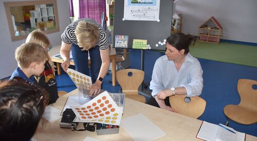 Zwei Kinder und drei Frauen am Tisch. Aufkleber von gelben und orangenen Smileys liegen auf dem Tisch. Frau mit kurzen blonden Haaren kramt noch mehr Smileys hervor. Frau mit schwarzen Haaren hockt am Tisch und stützt sich mit einem Arm auf Kinderstuhl.