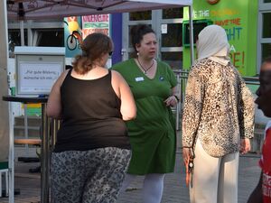 Zwei sehr korpulente Frauen unterhalten sich mit einer schlanken Frau mit dünner Jacke in Leoparden-Optik und hell-beigen Kopftusch. Es ist ein Thementisch auf einem Schild steht: Für gute Bildung wünsche ich mir....