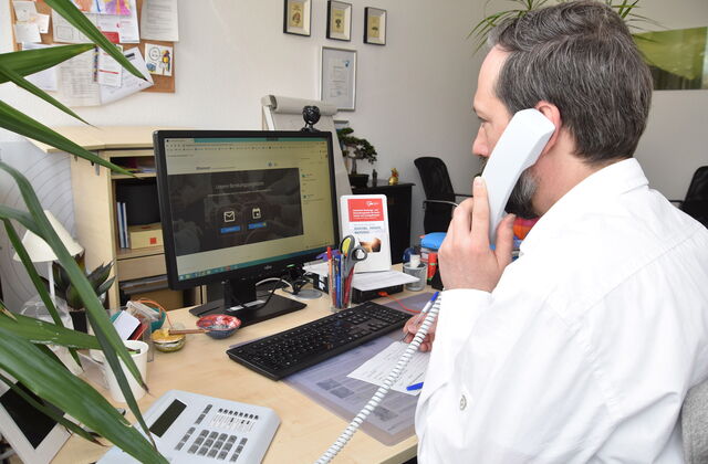 Ein Mann in weißem Hemd sitzt in einem NBüro am Schreibtisch, vor ihhm der Monitor, Tastatur unnd ein Notizblock, wo er etwas notiert, er hat den Telefonhörer am Ohr.