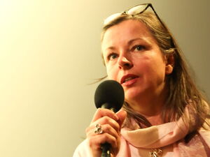 Frau mit Mikrofon in der Hand. Jung, lange Haare, Brille, helles kurzärmliges Oberteil und Schal/Tuch um den Hals.