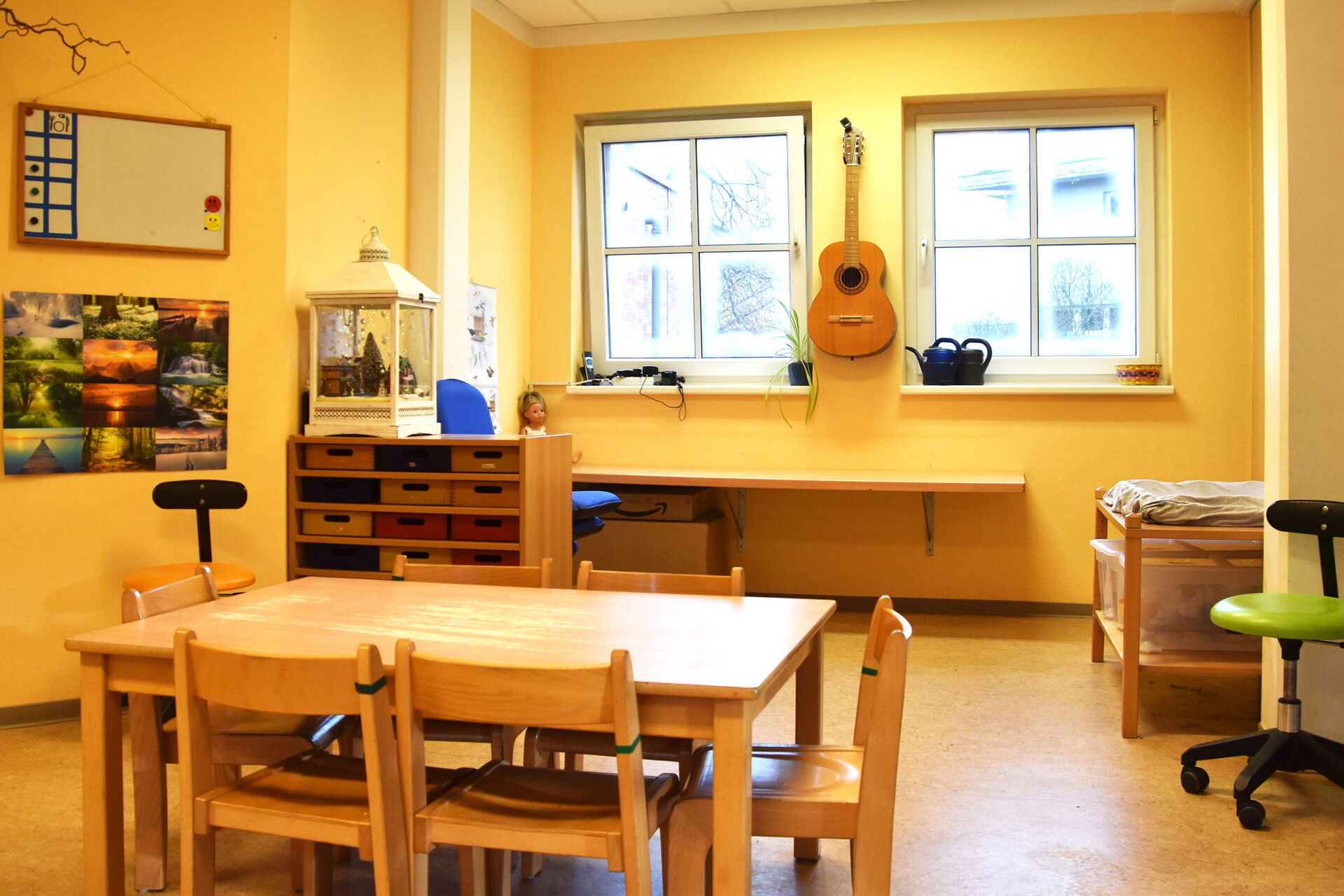 Bastelzimmer. Tisch mit 6 Stühlen. Gelbe Wände. Zwischen zwei Fenstern hängt eine Gitarre an der Wand.