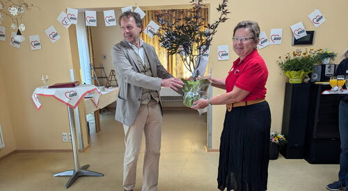 Großer Raum. Ein Stehtisch. Ein Mann übergibt eine Topfpflanze an eine Frau. Der Mann ist groß, krauses graue Haar, graues Jackett, weißes Hemd, helle Hose, braune Schuhe, Frau mit Brille, rotem AWO Poloshirt und schwarzen Rock, in Sandalen.